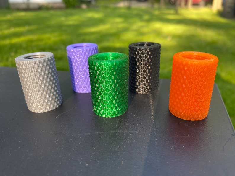 Puede incluir: Cinco objetos cil&iacute;ndricos de diferentes colores con una superficie texturizada. Los colores son gris, morado, verde, negro y naranja.