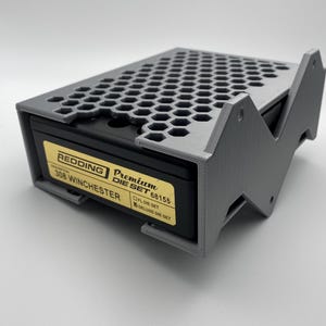 Puede incluir: Una caja de almacenamiento de plástico gris para matrices de recarga. La caja tiene un patrón de panal en la parte superior y una etiqueta que dice "REDDING Premium DIE SET 68155 308 WINCHESTER FL. DIE SET DELUXE DIE SET".