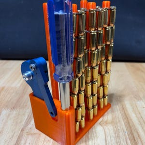 May include: An orange 3D-printed ammunition holder with a blue screwdriver and a blue tool. The holder contains numerous brass-colored bullets. The holder is on a wooden surface.