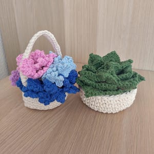 May include: Two crochet items: a basket with a handle filled with pink, blue, and purple flowers, and a small bowl with green leaves. Both are made of white and colored yarn, and sit on a wooden surface.
