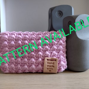 May include: A pink crocheted pouch with a brown leather tag that says "hand made with" and a heart. The pouch is sitting on a wooden surface next to two gray objects.