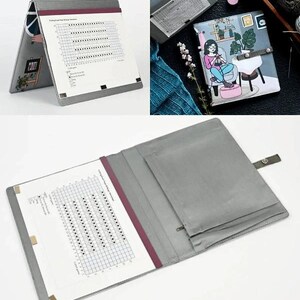 May include: A gray fabric pattern keeper folder featuring a graphic illustration of a person knitting in a chair. The interior includes a white grid chart, a magnetic closure strap, and a zippered pocket for organizing small crafting accessories.