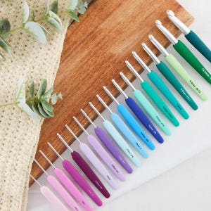 May include: A set of crochet hooks with ergonomic handles in a gradient of colors ranging from light pink to dark teal. Each hook features a silver metal tip and is clearly labeled with its specific size in millimeters and US letter sizing.