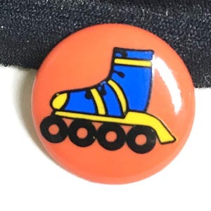 May include: A circular pin-back button featuring a vibrant orange background with a graphic illustration of a blue inline skate. The skate has yellow accents on the boot and frame, and four black wheels positioned along the bottom edge.