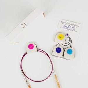 May include: A collection of knitting accessories, including circular needles with a 24-inch (61 cm) cord, and Snap ID cord organizer tags in various sizes. A white box with the number "21" is also present.