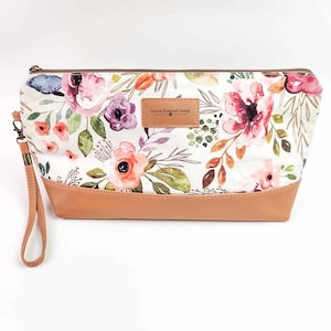 May include: A floral print cosmetic bag with a tan leather base and wrist strap. The bag features a white background with pink, purple, and orange flowers. The text "twice sheared sheep" is on a small leather tag.