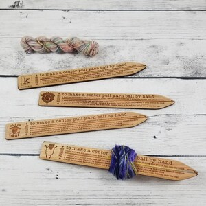 May include: Four light-colored wooden yarn winding sticks with engraved instructions for making a center pull yarn ball. Each stick features a unique logo and a notch. One stick has a small ball of purple and green yarn wrapped around it.
