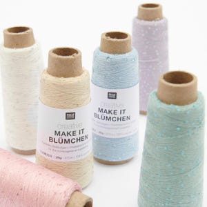 May include: Five spools of Rico Design Creative Make It Blümchen yarn in pastel shades of cream, light blue, lavender, pink, and mint green. Each spool features a cardboard core, a white paper label with black text, and a subtle metallic sequin finish.