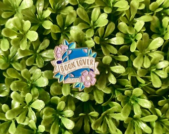 May include: Gold enamel pin with a blue book, pink flowers, and green leaves. The book has the text "BOOK LOVER" on it.