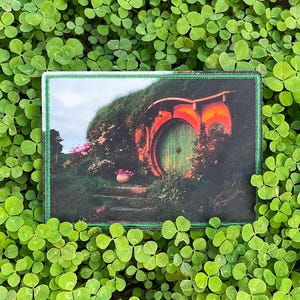 May include: A rectangular patch featuring a hobbit hole with a green door, orange accents, and a grassy roof. The patch is surrounded by a green border and set against a backdrop of green clover.
