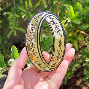 May include: A gold-colored ring with a reflective surface and black engraved script lettering wrapping around the entire outer band. The ring features a distinct oval shape and is held up against a background of green leaves and natural sunlight.