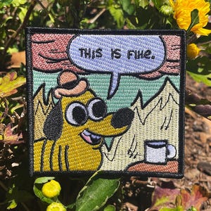 May include: A square embroidered patch featuring a yellow dog wearing a brown hat, sitting at a table with a white coffee mug. The background shows flames, and a speech bubble above the dog contains the black text, THIS IS FINE.