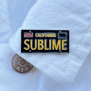 May include: A black and yellow enamel pin with a California license plate design. The pin says "Sublime" and "Long Beach" in red letters.