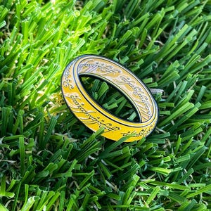 May include: A gold-colored metal ring featuring a black interior and black engraved script lettering that wraps around the entire outer surface of the band. The ring has a smooth, polished finish and a rounded profile, displaying intricate text.