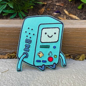 May include: A light blue embroidered patch of a cartoon robot with a smiling face, a screen that says "BMO", and a game controller on the front. The patch is sitting on a brown wooden surface.