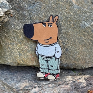 May include: An embroidered patch featuring a cartoon character with a tan head, a large black nose, and a light blue sweater. The character wears light green pants and red and white sneakers, with hands tucked into the pockets of the sweater.