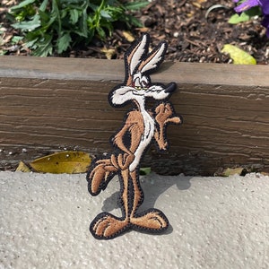 May include: A brown and white cartoon coyote embroidered patch with a sly expression. The patch is on a brown wooden surface with a flower bed in the background.