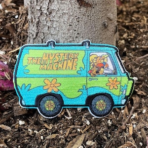May include: A green and blue embroidered patch of the Mystery Machine van from Scooby-Doo. The van has the words "Mystery Machine" on the side and features the characters Scooby-Doo and Shaggy driving.