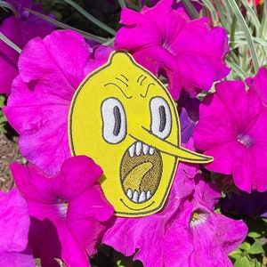May include: A yellow lemon-shaped patch with a cartoon face, an open mouth, and a long nose. The patch is placed on a bed of bright pink flowers.