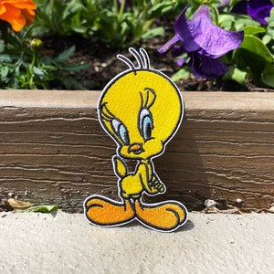 May include: A yellow Tweety Bird patch with black and white details and orange feet. The patch is embroidered with a black outline. The cartoon character is standing on a concrete surface, with flowers in the background.