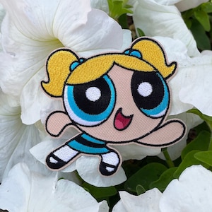 May include: A cartoon character patch of a girl with blonde pigtails, blue eyes, and a pink face. She is wearing a blue and white striped dress and white socks. The patch is surrounded by white flowers.