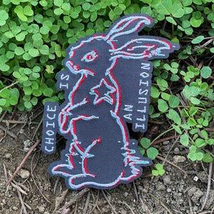 Peut inclure: Un écusson gris foncé représentant un lapin avec des contours rouges et bleus. Le texte "CHOICE IS AN ILLUSION" est en blanc. L'écusson est posé sur un lit de trèfles verts.