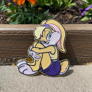 Peut inclure: Un patch brodé représentant un lapin de dessin animé assis, les jambes croisées. Le lapin porte une tenue violette et jaune.