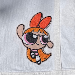 May include: An orange and pink embroidered patch of a cartoon girl with red hair and a white and black striped shirt.