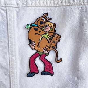 May include: A brown and orange cartoon dog with a green collar is hugging a blonde cartoon person wearing a green shirt and brown pants. The cartoon characters are embroidered on a white fabric.