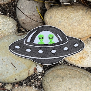 May include: A black and white embroidered patch of a UFO with two green aliens inside. The patch is on a bed of rocks.