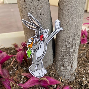 May include: An embroidered patch of Bugs Bunny holding a carrot, with a red nose and white eyes. The patch is attached to a tree trunk.