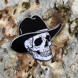 May include: A black and white embroidered patch of a skull wearing a cowboy hat. The skull is white with black eyes and a black outline. The hat is black with a white band.