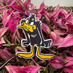 May include: Black and yellow cartoon duck character with a grumpy expression. The duck is standing on a bed of pink and green leaves.