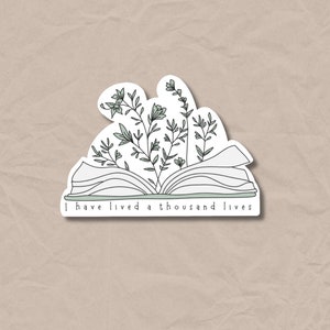 Puede incluir: Una pegatina con un libro blanco abierto con plantas verdes que crecen de él. El texto "I have lived a thousand lives" está escrito debajo del libro.