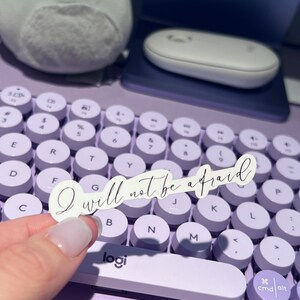 以下が含まれることがあります： 丸いキーを持つ紫色のキーボードに、「I will not be afraid」というテキストが書かれた白いステッカーが貼られています。キーボードには、「logi」と印刷された銀色のスペースバーがあります。
