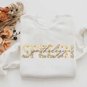 Puede incluir: Sudadera blanca con las palabras "Speech Pathology" en letras florales. Las letras están hechas de flores amarillas y naranjas con hojas verdes.