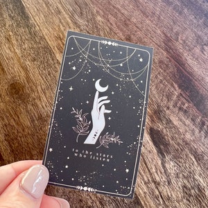 K&ouml;nnte beinhalten: Eine schwarze Karte mit einer wei&szlig;en Hand, die zu einem Halbmond greift. Der Hintergrund ist dunkelblau mit wei&szlig;en Sternen und einem wei&szlig;en kettenartigen Design. Der Text "to the stars who listen" ist in wei&szlig; am unteren Rand der Karte gedruckt.