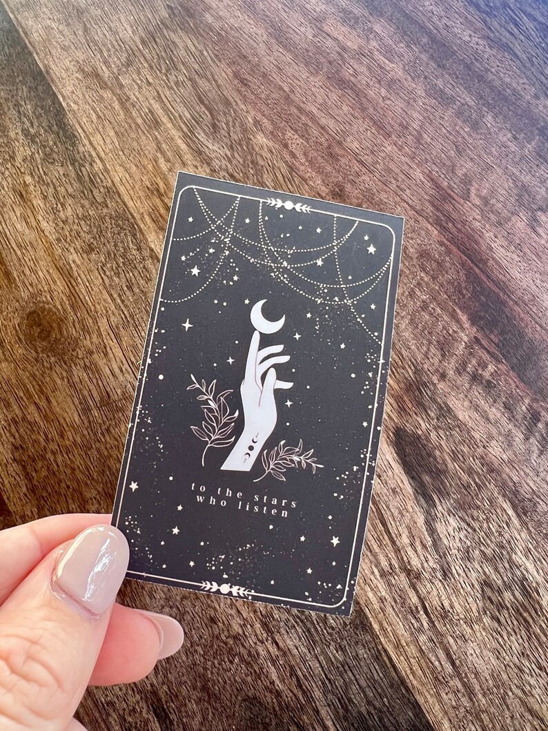K&ouml;nnte beinhalten: Eine schwarze Karte mit einer wei&szlig;en Hand, die zu einem Halbmond greift. Der Hintergrund ist dunkelblau mit wei&szlig;en Sternen und einem wei&szlig;en kettenartigen Design. Der Text "to the stars who listen" ist in wei&szlig; am unteren Rand der Karte gedruckt.
