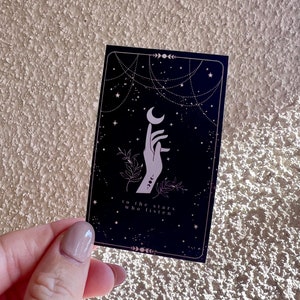 K&ouml;nnte beinhalten: Eine schwarze Karte mit einer wei&szlig;en Hand, die nach einem Halbmond greift. Der Hintergrund ist dunkelblau mit wei&szlig;en Sternen und einem wei&szlig;en kettenartigen Design. Der Text auf der Karte lautet "to the stars who listen".