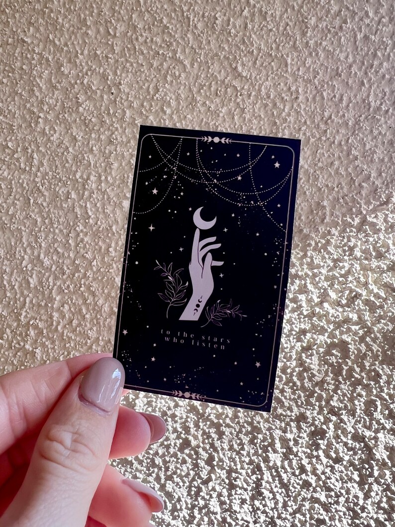 K&ouml;nnte beinhalten: Eine schwarze Karte mit einer wei&szlig;en Hand, die nach einem Halbmond greift. Der Hintergrund ist dunkelblau mit wei&szlig;en Sternen und einem wei&szlig;en kettenartigen Design. Der Text auf der Karte lautet "to the stars who listen".