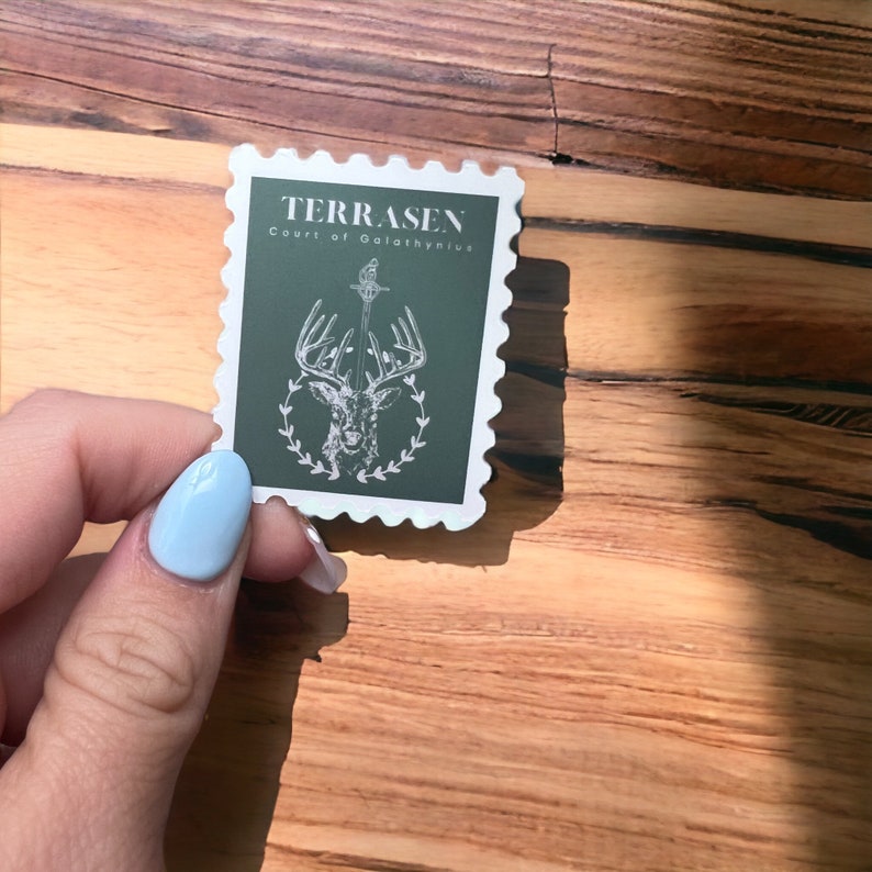 May include: A white sticker with a green background and a white outline. The sticker features a white stag with antlers and a sword, surrounded by a wreath of leaves. The text "TERRASEN Court of Galathynius" is printed in white at the top of the sticker.