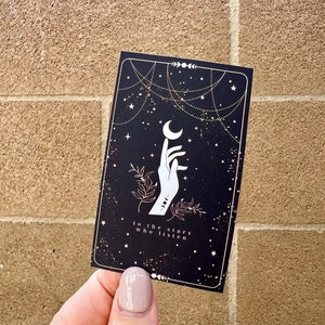 K&ouml;nnte beinhalten: Eine schwarze Karte mit einer wei&szlig;en Hand, die nach einem Halbmond greift. Der Hintergrund ist eine schwarze, sternen&uuml;bers&auml;te Nacht mit goldenen Sternen und einer goldenen Kette. Der Text auf der Karte lautet "to the stars who listen".