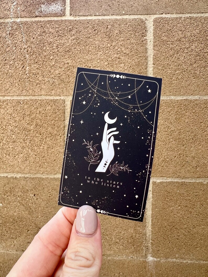 K&ouml;nnte beinhalten: Eine schwarze Karte mit einer wei&szlig;en Hand, die nach einem Halbmond greift. Der Hintergrund ist eine schwarze, sternen&uuml;bers&auml;te Nacht mit goldenen Sternen und einer goldenen Kette. Der Text auf der Karte lautet "to the stars who listen".