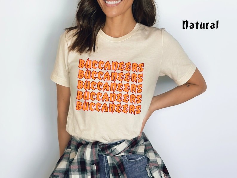 May include: Cream-colored t-shirt with the word "BUCCANEERS" in orange block letters repeated vertically. The word "Natural" is printed in black script in the upper right corner. The person is wearing a plaid shirt tied around the waist.