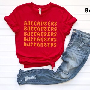 May include: A red t-shirt with the word "BUCCANEERS" printed in orange multiple times. The shirt is paired with blue denim jeans. The word "Red" is written in the upper right corner.