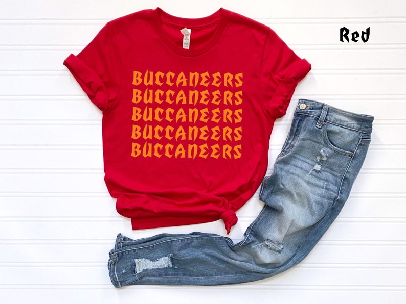 May include: A red t-shirt with the word "BUCCANEERS" printed in orange multiple times. The shirt is paired with blue denim jeans. The word "Red" is written in the upper right corner.