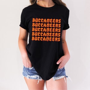 May include: Black t-shirt with the word "BUCCANEERS" printed in orange, repeated five times. The text has a bold, stylized font. The shirt is a crew neck style.