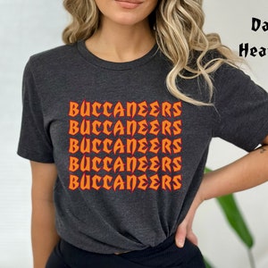 May include: Dark heather gray t-shirt with the word "BUCCANEERS" in orange, repeated five times. The text has a stylized, bold font. The shirt is short-sleeved and appears to be made of a soft material.