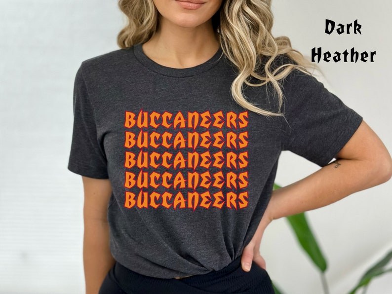 May include: Dark heather gray t-shirt with the word "BUCCANEERS" in orange, repeated five times. The text has a stylized, bold font. The shirt is short-sleeved and appears to be made of a soft material.
