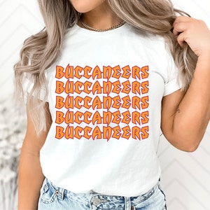 May include: White t-shirt with the word "BUCCANEERS" in orange and red repeating across the front. The text has a bold, slightly distressed design. The shirt has short sleeves and a crew neck.